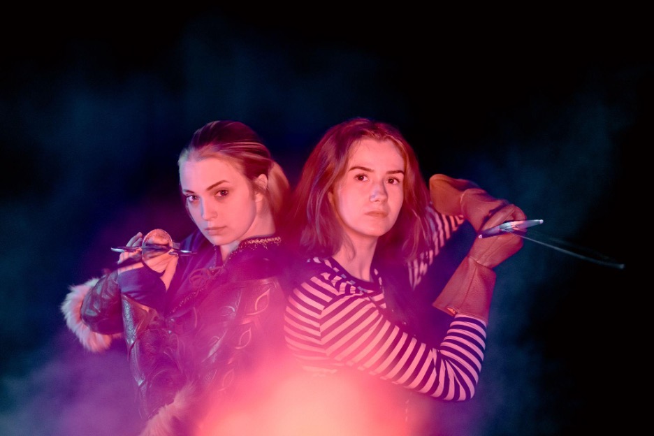 Two young women, the one on the left in traditional fantasy clothing and the one on the right in modern day clothing, hold swords up, pointed towards the camera. They are in a dark room, with purple and pink colored smoke.