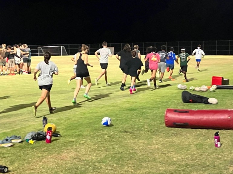 Athletes are running together on a grass field at night. Sports equiptment are scattered on the ground.