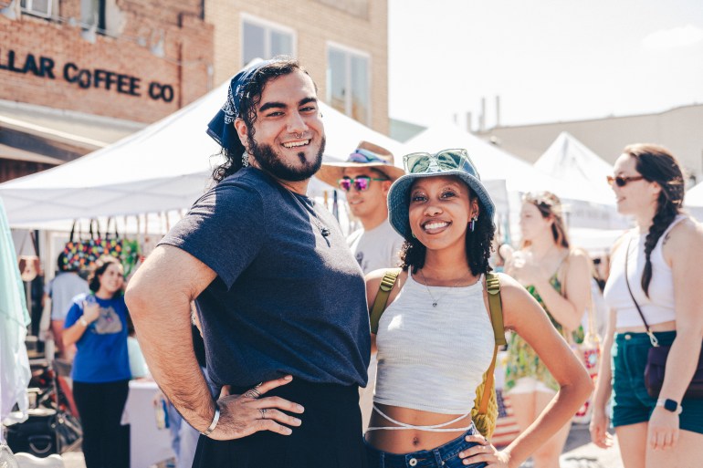 Two people standing side by side in center focus. One on the left has a dark blue bandana on their head and the one on the right has a light-blue jean bucket hat with sunglasses on top. The person on the left has facial hair and a septum piercing, and they are wearing a dark blue t-shirt. The girl on the right is wearing a white halter tank top and green backpack straps showing. The background is out of focus with people passing by in front of a coffee shop with white vendor tents.
