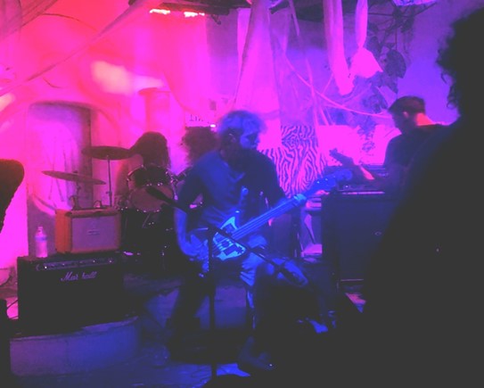 On stage is Kromathaw, bassist Issac King is in the middle illuminated by pink and blue lights, in the background on drums is Sol Jäger and guitarist Clark Drake, while lead singer Rowan Latta is on the floor singing into the microphone.