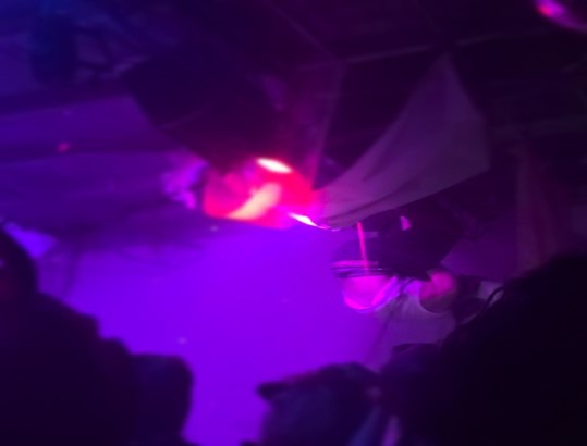The image shows lead singer of Kromathaw, Rowan Latta, hanging upside down from the rafters of the music venue, The Electric Church, while wearing a pope’s hat as the pink and purple lighting illuminates the performance.