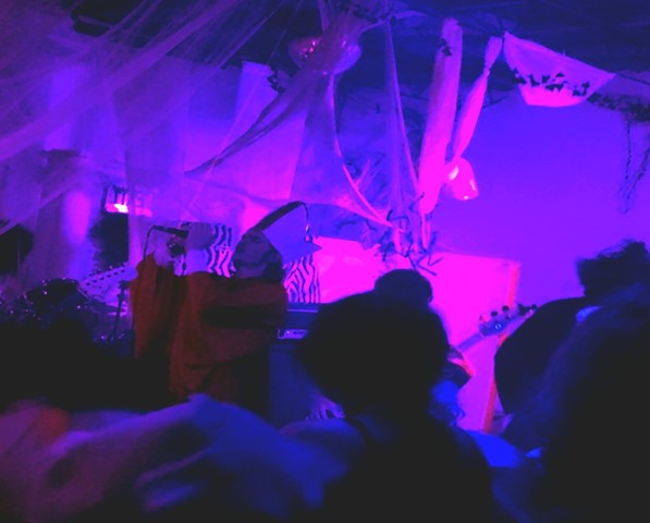 In the photo Kromathaw is performing in front of a crowd, the lead singer, Rowan Latta, is singing into the microphone while wearing a pope costume, while bassist Issac King is strumming away next to him