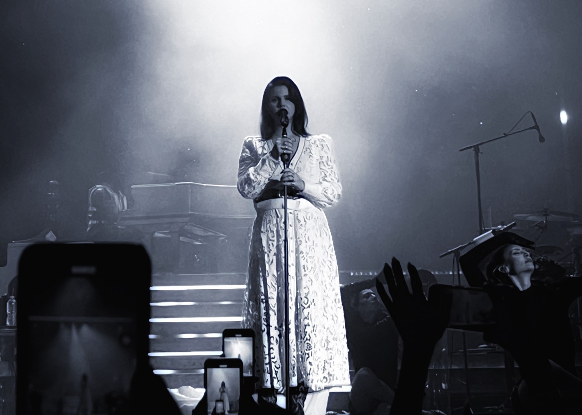 Photo includes Lana Del Rey on stage wearing white dress and black belt while she is singing.