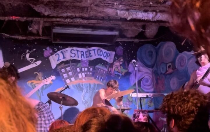 The image shows a man playing on a drumset with a mic also in front of him. Behind him is a wall with an art design. Included on the art design is a house and the text “21st Street Co-op”.
