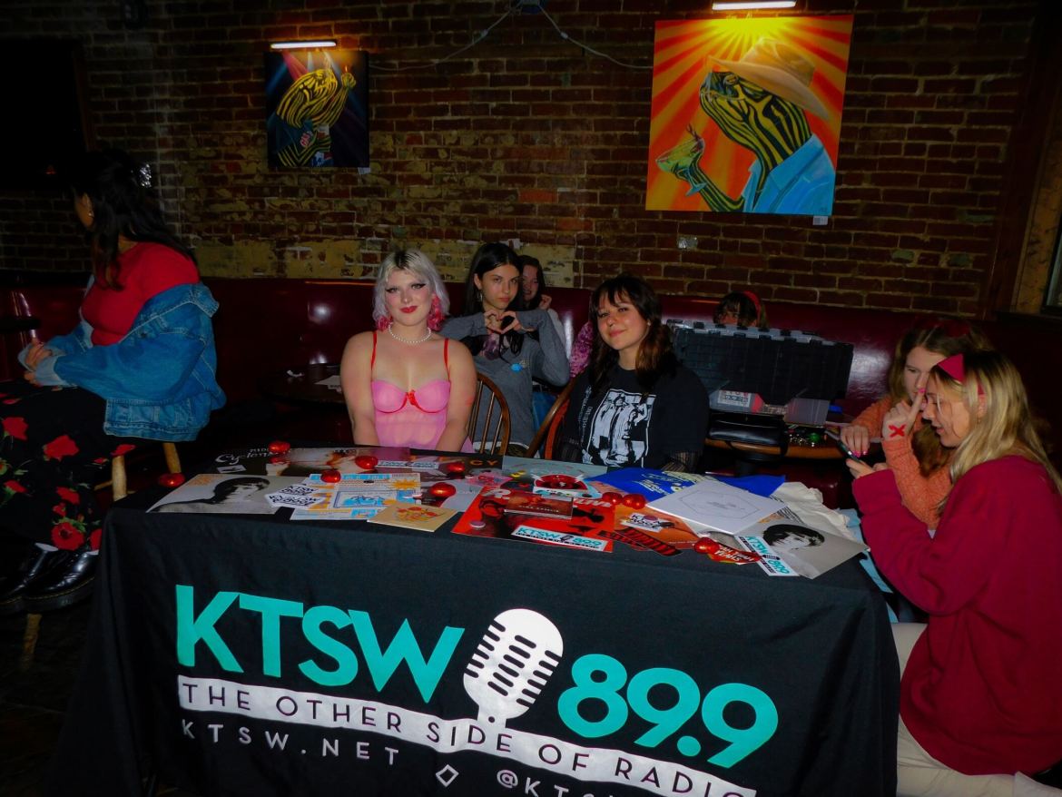 The image shows three women sitting at a table. The table has a large tablecloth that says “KTSW 89.9” “The Other side of radio” “KTSW.net”. There is also an image of a microphone on the tablecloth. There are two women to the right of the table and one woman to the left. The background of the image is a brick wall, and on the wall are two paintings of lizards in clothes.