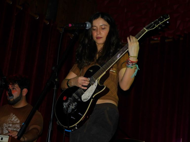 The image shows a woman playing a guitar with a mic also in front of her. To the woman’s left is a man’s face with a mic in front of his face. 