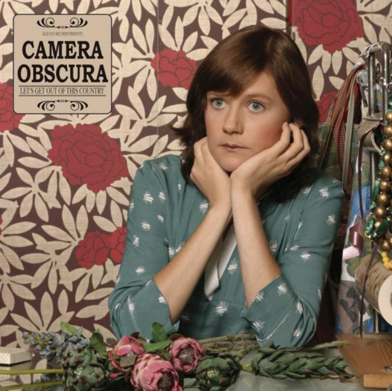 Tracyanne Campbell wearing a flowy teal top with her arms resting on a table and her hands on her face. She is in front of red flower wallpaper and flowers on the table in front of her. An old-looking logo with Camera Obscura and the album title is in the top left corner.