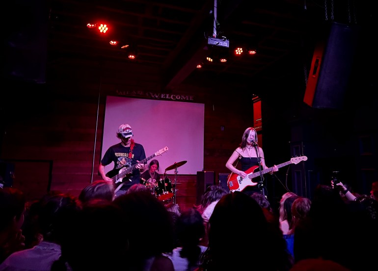 The band Pretty Sick performing in front of a medium-sized crowd. Red lights shine down. 