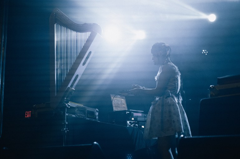 Right side perspective of Alice Longyu Gao (ALG) with concert light shining on them. ALG’s pink harp is on the left of the picture and Alice is adjusting their equipment with their computer in the background.