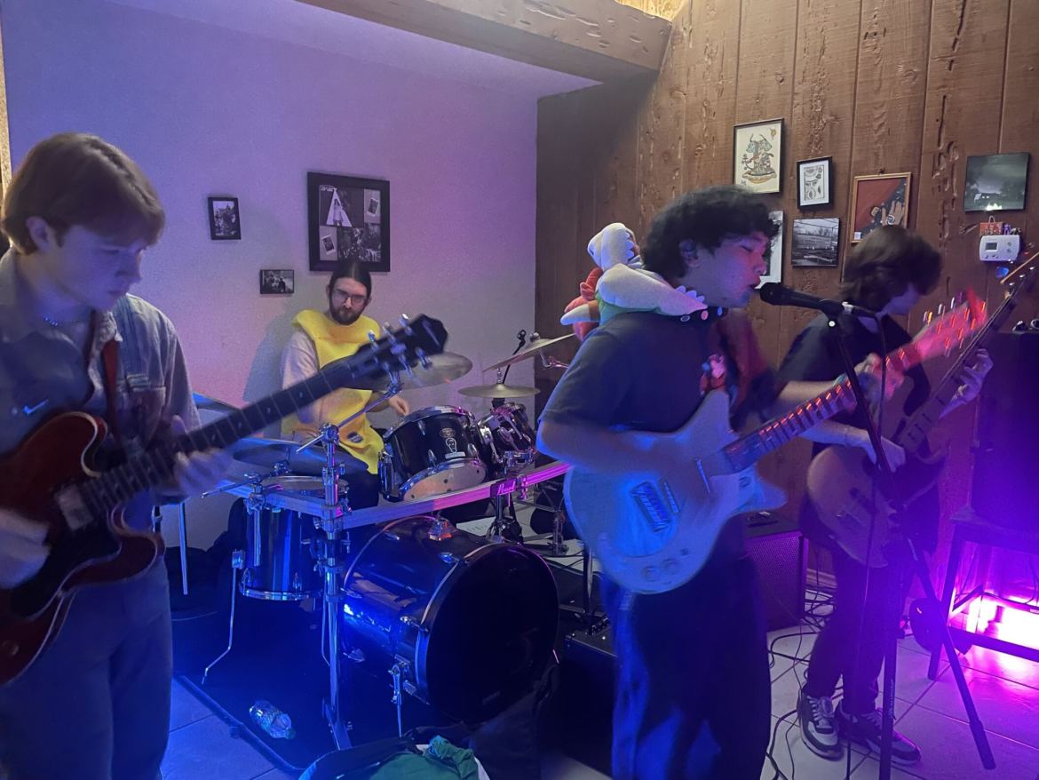 The image shows four people. On the left is a man with a guitar. In the middle-back is a man in a banana costume who is sitting at a drum set. Slightly askew from the middle, in the front is a man with a guitar and a microphone in front of him. On the right is another band with a guitar. The lighting of the photo is purple and blue.