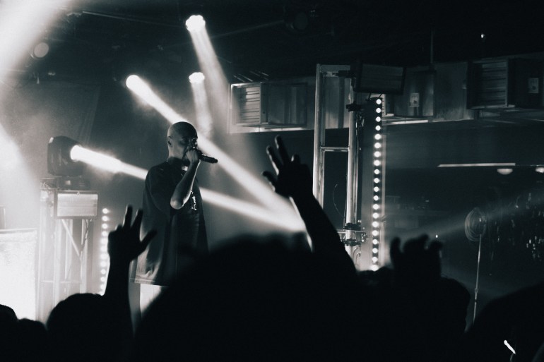 Danny Brown holding a mic. The shot has the hands of fans reaching towards him as he looks into the audience. It is in dark blue and white hues.