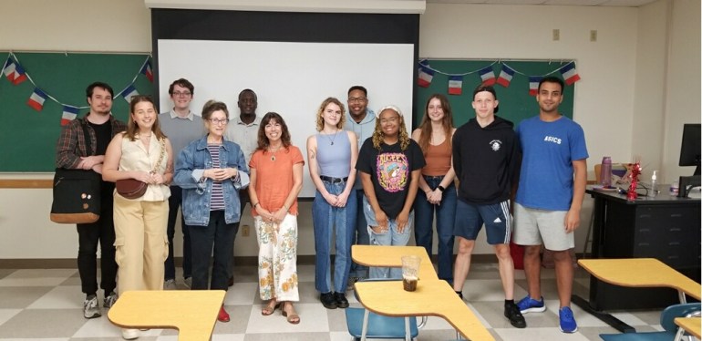 12 members of the French Club standing in front of a white projector and a green chalk bored with small French flags hanging on the border of the green chalk board. There are 3 desks presented in the photo.