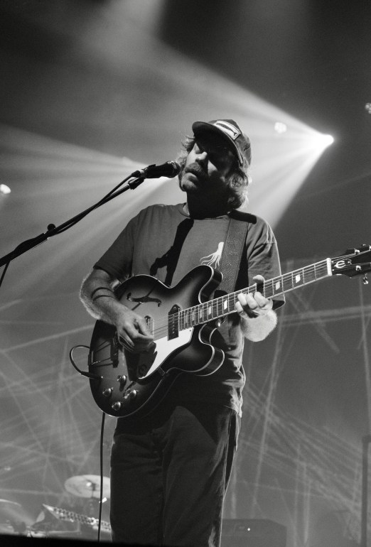 Portrait photo of the guitarist from slowdive in a cap with a fish on it, dark t-shirt, and jeans while singing on the microphone and playing the guitar.