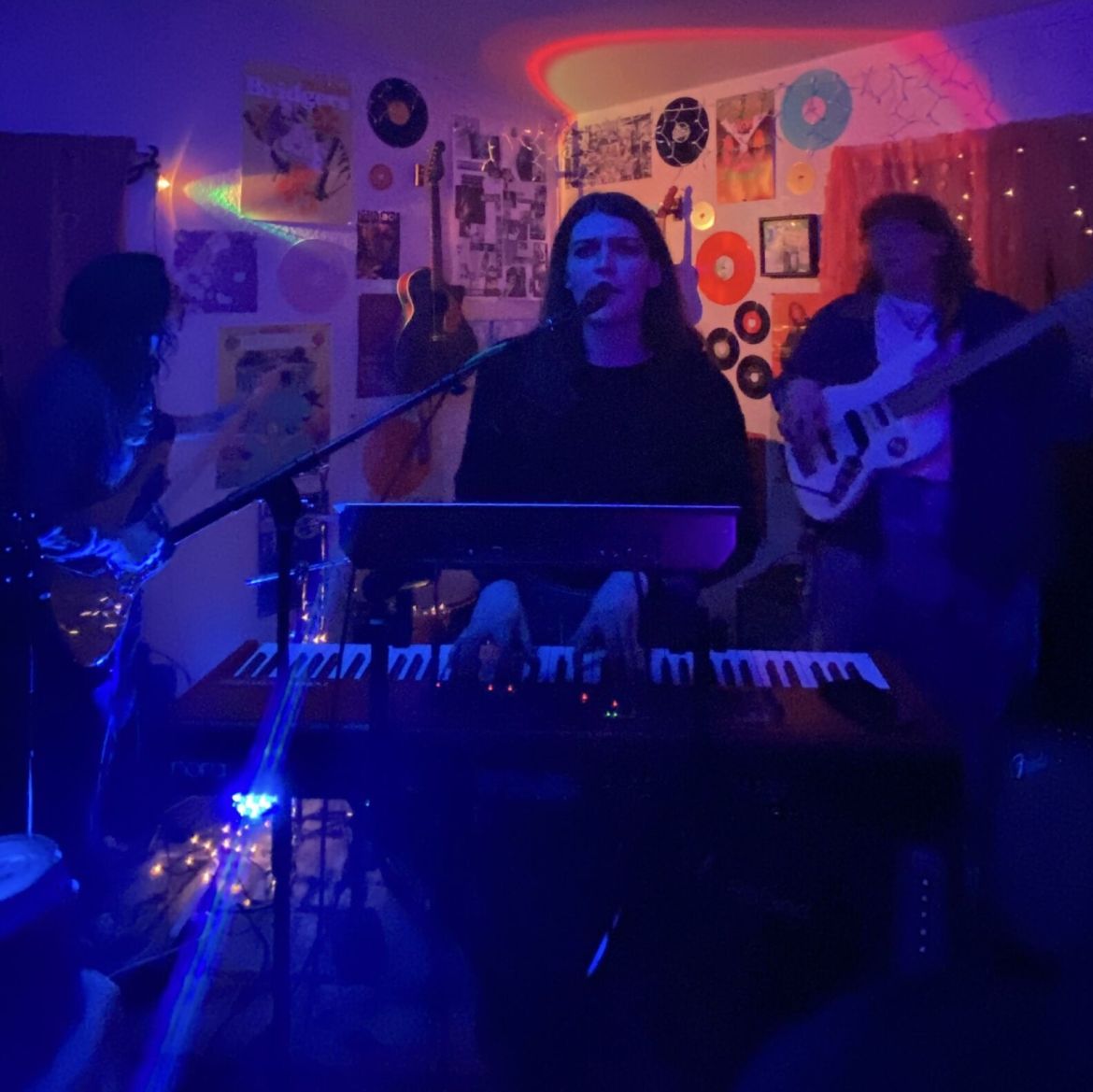 The image shows three people. On the left is a person with a guitar. In the middle is a person who is standing at a keyboard with a microphone in front. On the right is another person with a guitar. The lighting of the photo is blue with an orange background that has posters, vinyl’s, and a guitar hung on the wall.