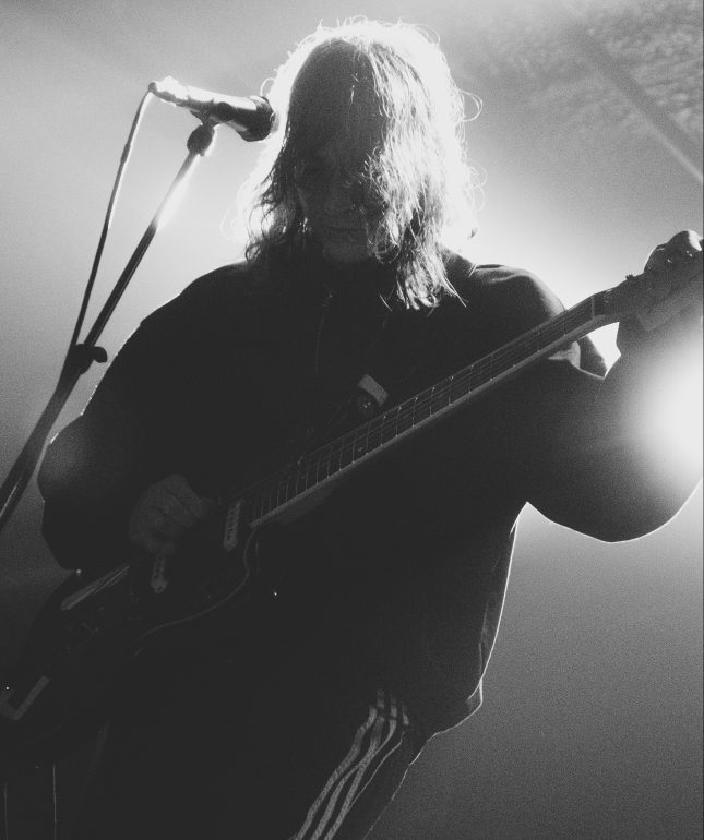 Mk.gee in black and white. Microphone is on the left side of his face and he is looking down. His bangs cover his face and not much is able to be made out since the lighting is from behind.