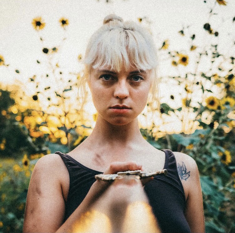 girl with blond hair pointing a sword to the camera in front of sunset and sunflowers