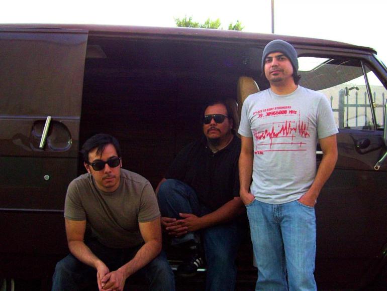 Nic Gonzales, Jason Gonzales, and Matt Anderson are pictured from left to right in front of a brown tour van. Nic and Jason are seated wearing sunglasses, while Matt who’s standing, wears a beanie. All three are wearing blue jeans and t-shirts, while Matt wears “A Place to Bury Strangers” graphic tee. 