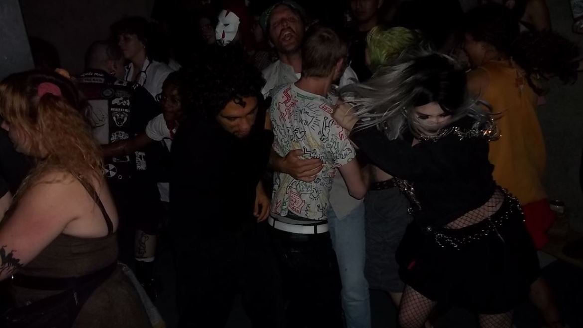 A group of people taking part in a mosh pit at a Bosh show. The image is taken in the action, the people photographed moshing in a dark garage. 