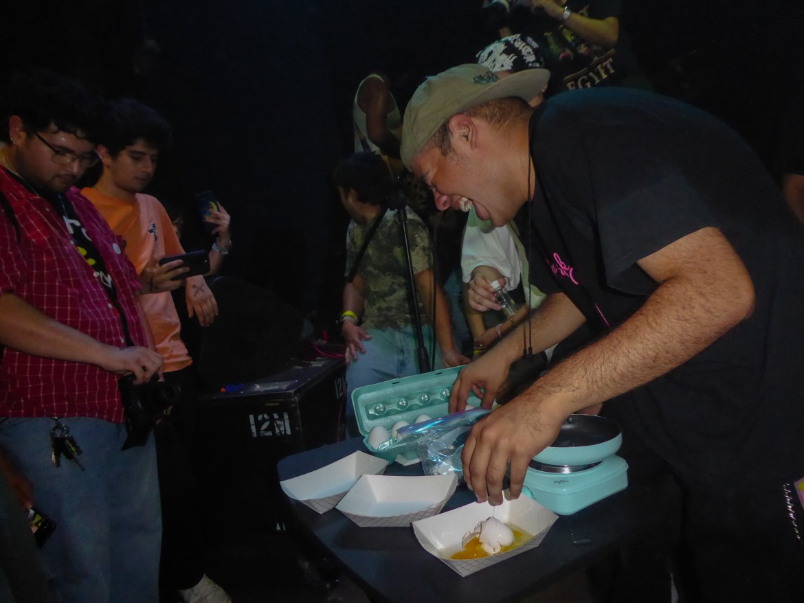 Man laughing as he is making scrambled eggs at a small table surrounded by people taking photos in a dark room