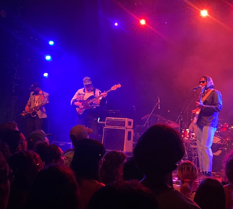 3 people on a blue and red lit stage with guitars and crowd in front