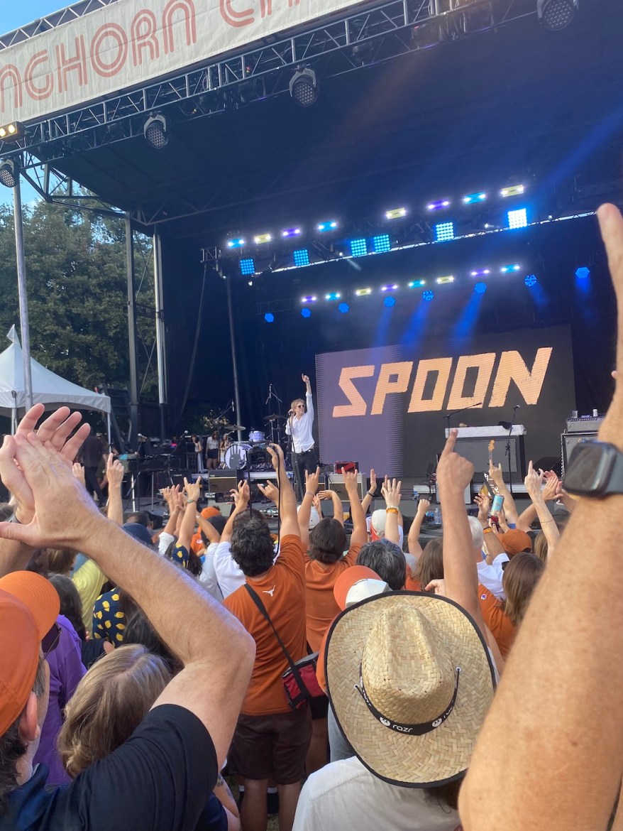 band singing in front of crowd wearing burnt orange longhorn merch cheering at a a black stage with blue lights