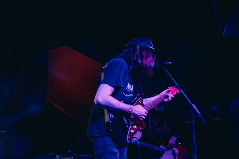 Photo of Aaron Powell (Fog Lake) playing guitar and singing into the mic. Purple lighting overcasts him.
