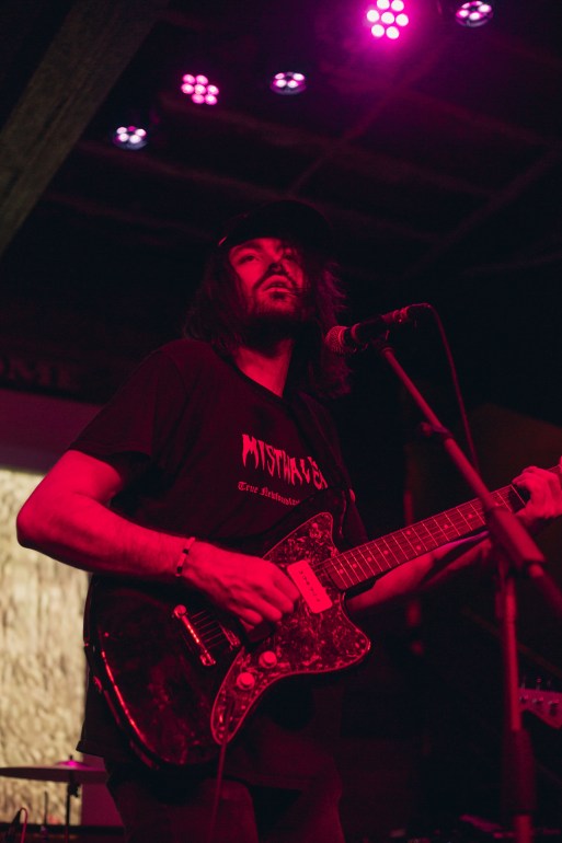 Aarron Powell (Fog Lake) looks into the crowd while strumming his guitar. The lighting is red. 