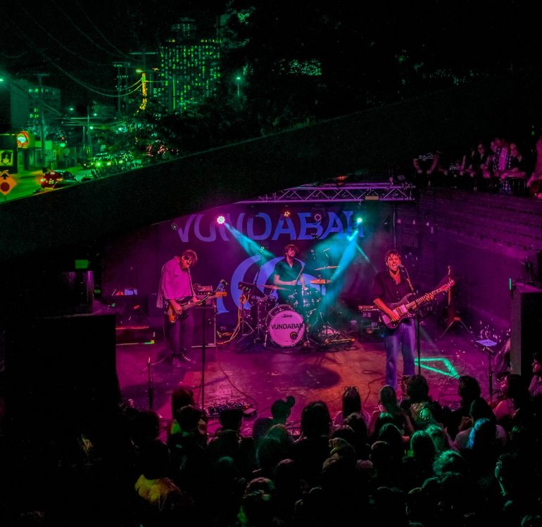 Vundabar performs live at Mohawk in Austin, Texas. The lead singer stands center stage, guitar in hand, as vibrant lights illuminate the band. The packed crowd is energized, with hands raised, fully immersed in the music