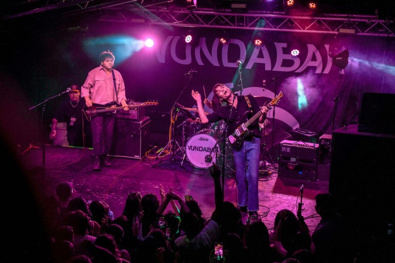 Pink lit stage with word Vundabar behind two guitarists and drummer performing