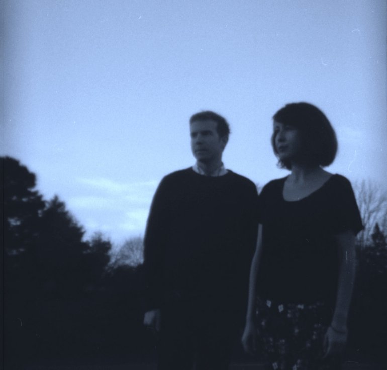 A pale dark blue photograph of Alice Hansen and Chuck Blazevic from You’ll Never Get To Heaven outside facing off into the distance with black trees behind them.