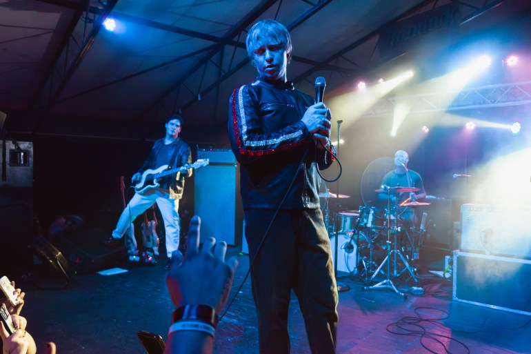 The Drums band on blue stage with lights flashing around. Lead singer is looking down at the crowd with a sassy face. 