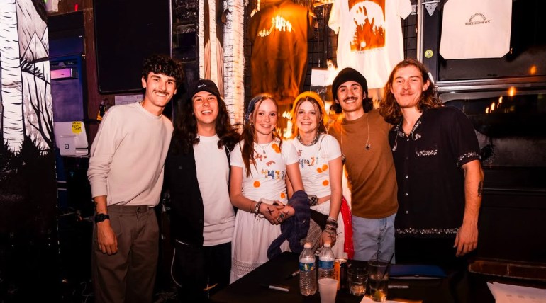 Nick, Alan, Tomas, and Cogan are standing in front of the merch table. Between them are two fans wearing white t-shirts with “The 4411” painted on them in bright colors. Behind them, there are two shirts and a tote bag, displaying the available merchandise. In front of the group, there is a black table, holding water bottles and water cups. 