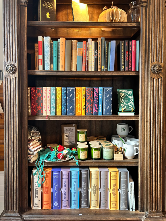 Candles, mugs, crochet gifts, board games, and novels fill a brown bookshelf at the Green Heron Bookshop.