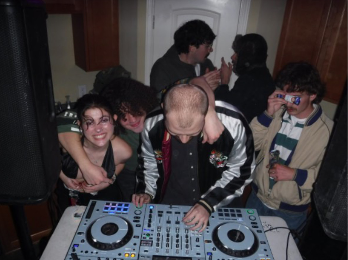 The image contains a DJ working a large grey turntable, Hannah’s friend has one arm around the DJ and another around Hannah, the two are smiling at the camera. On either side of the image, there are large speakers. In the background, two men are talking to each other. On the other side of the DJ, holding a granola bar over his eyes.