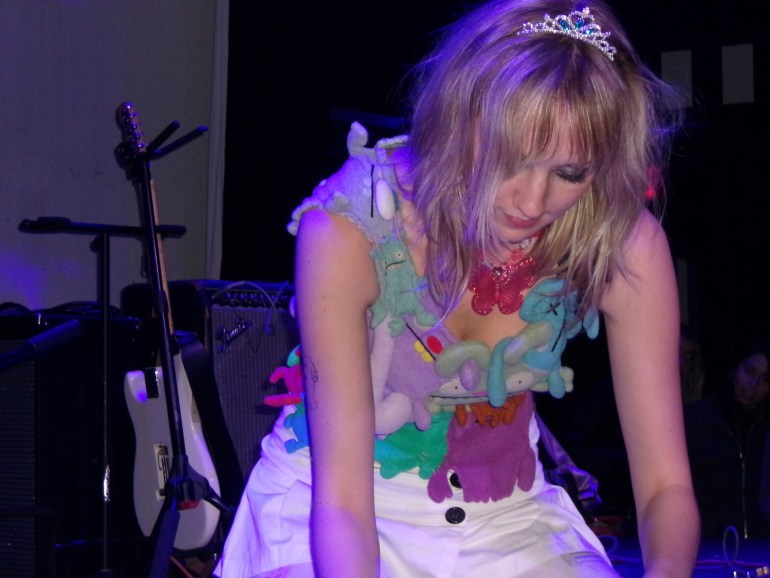 Photo of Lydia Slocum of Feeble Little Horse on stage at the Parish in Austin. She is knelt down on the ground messing with equipment. She is wearing her handmade Ugly Doll top, a white skirt, a plastic pink butterfly necklace and a tiara.