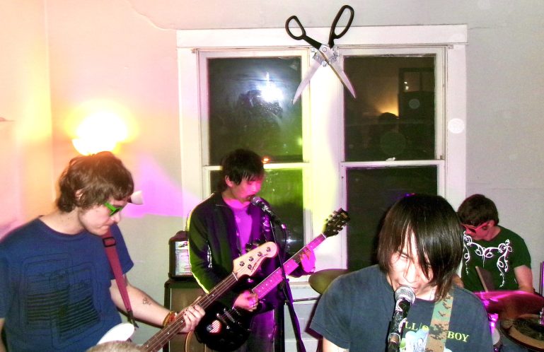 Photo of Push Ups at Scissor House. Theres pink and green-ish lighting. A cardboard cutout of scissors is hung up on the window behind the band