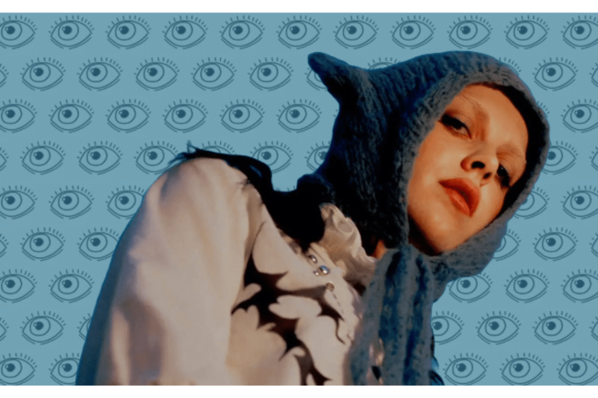 The picture is a photo of Ally Evenson wearing her signature blue crocheted hat. The background is blue with a bunch of drawn eyeballs.