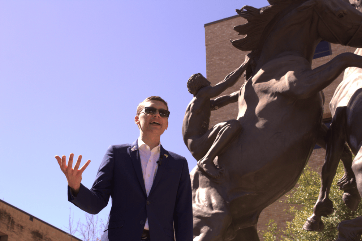 President of a student group makes a speech with a stallion statue behind him.