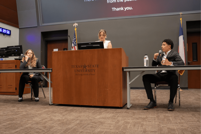 One candidate speaks into a microphone while the candidate to the left and the moderator listen. 