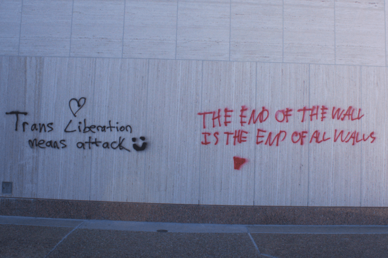 Trans liberation mean attack" with a heart and smiley face, and "The end of the wall is the end of all walls" with a red upside-down triangle." painted on the walls outside of Alkek library.