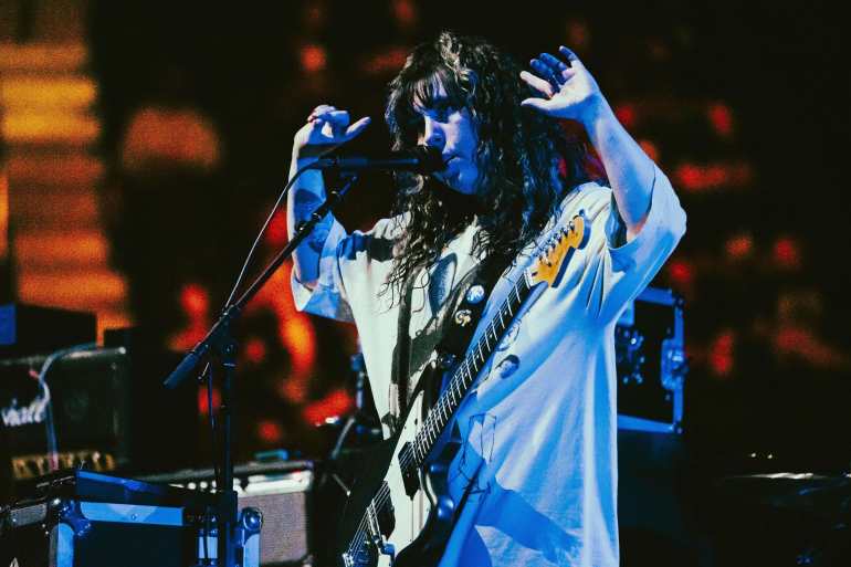 A woman with a black and white guitar has her hands up with a bright light shining down on her as she is surrounded by a crowd. 