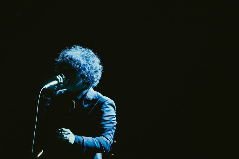 a man swallowed in darkness with nothing but a small light shining on his back as he holds a microphone and sings to the audience 