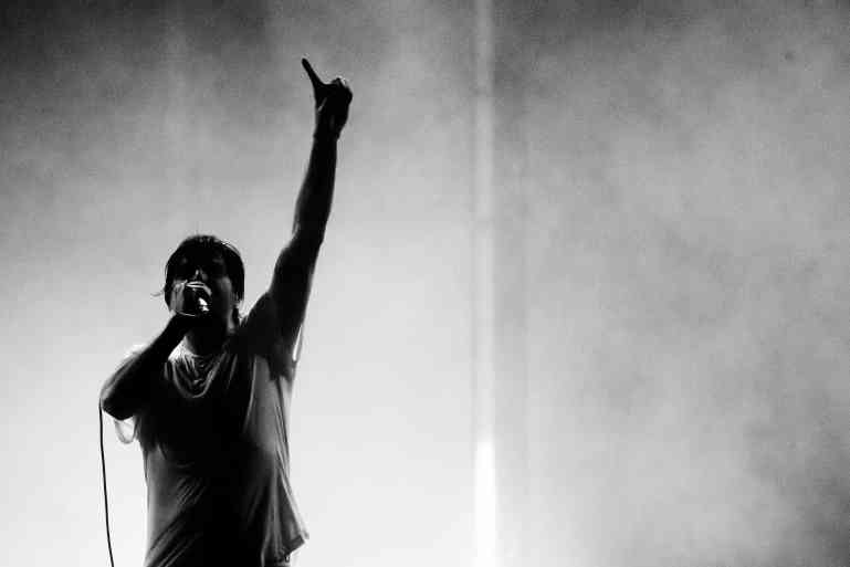 a man is standing on the bottom left-hand side holding a microphone in his right hand and his left hand up with finger pointed to the sky, illuminated in black and white with the light casting a light silhouette on the man's figure.