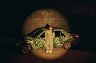 Samara Cyn poses in front of a green wrapped car with eyeball designs doing a photoshoot for her 2024 album, “The Drive Home”. She is lit by a single spotlight.