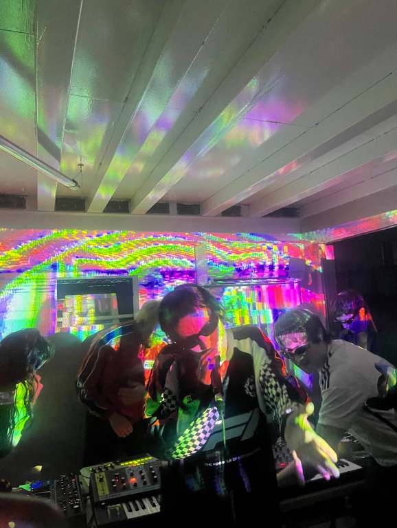 Pictured are five out of six members of the opera operating on MIDI keyboards and a mixer board while all wearing black shades with multicolored lighting covering them with one female in the front wearing a blue checkered F1 racing shirt holding a microphone