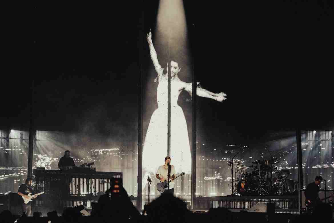 Five men pictured on stage, the two on the left with one playing the bass and the other playing the electronics, in the center is Chino Moreno holding a guitar with a mic in front and on the left side you have Abe Cunningham on the drums and another guitarist. A light shine on Chino in the middle with the picture of a women in a white dress dancing on the screen presented behind the band.