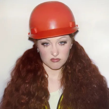 Chappell roan is pictured with long, voluminous red hair while wearing a bright orange construction helmet and a yellow safety vest over a white top. She has a serious expression with slightly pursed lips and a direct gaze at the camera. The background is a plain, off-white color. 