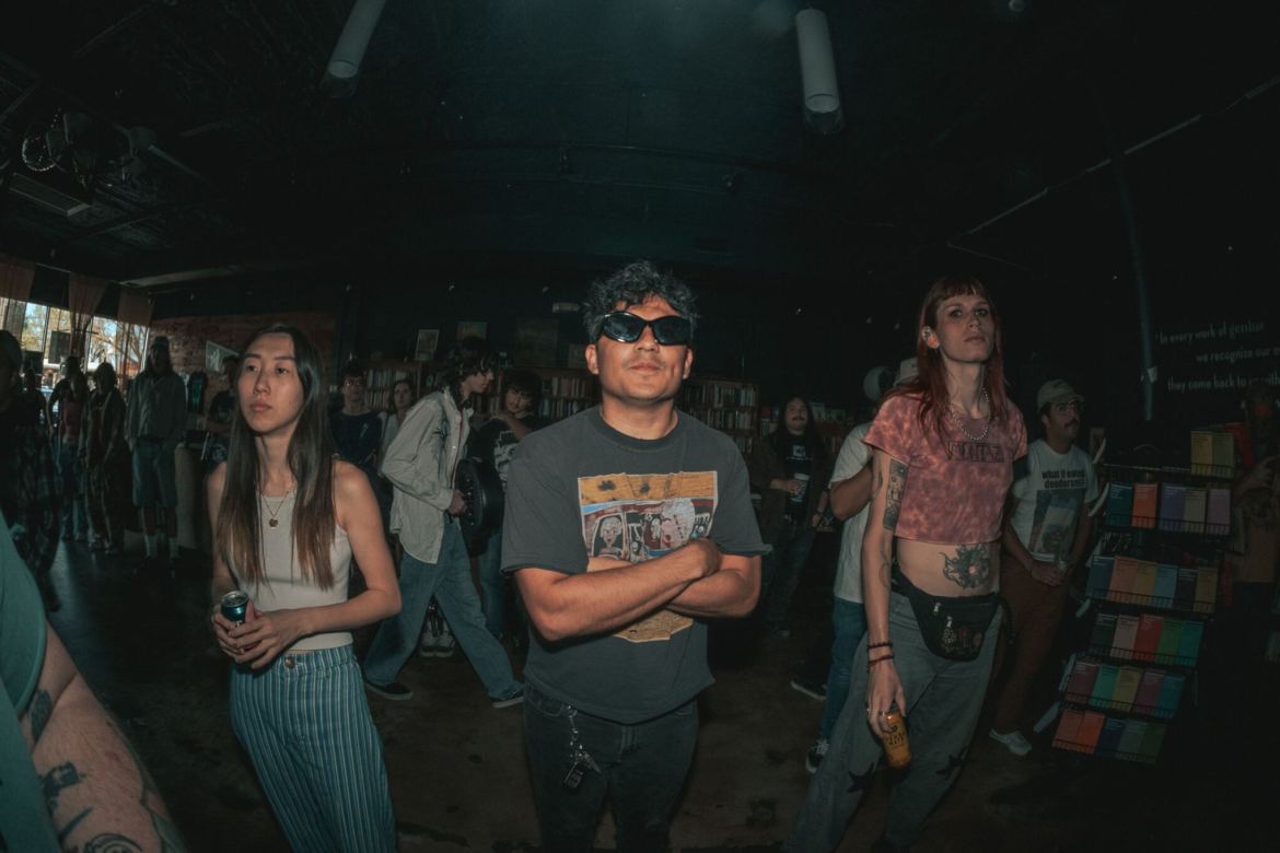 Taken with a fish-eye lens, the photo depicts three people in the foreground, one woman wearing a white tank top and blue and white striped pants, another individual in a pink patterned crop top, black fanny pack, and jeans. In the middle is Mat Zuniga with his arms crossed, sporting black sunglasses, a grey t-shirt, and black jeans. In the background, there is a crowd of people and a significant number of bookshelves.