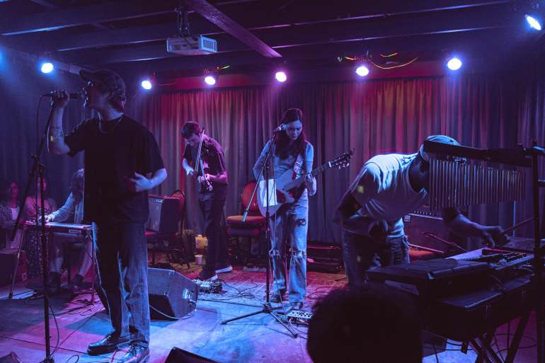 Chanel Beads stands together on stage in respective areas at their own instruments. To the very left is Mari Maurice on steel guitar; then Shane Lavers stands tall singing into the microphone, Zachary Paul drops his shoulder to play violin, Mya looks down while strumming her guitar, and leans over the pads playing beats for the set.