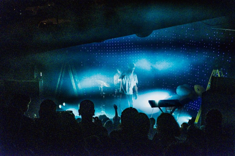Middle shot of George Clanton on stage with his drummer, Anya Good on the back of the stage to the left behind George. The backdrop is riddled with speckles of light that illuminated their figures.